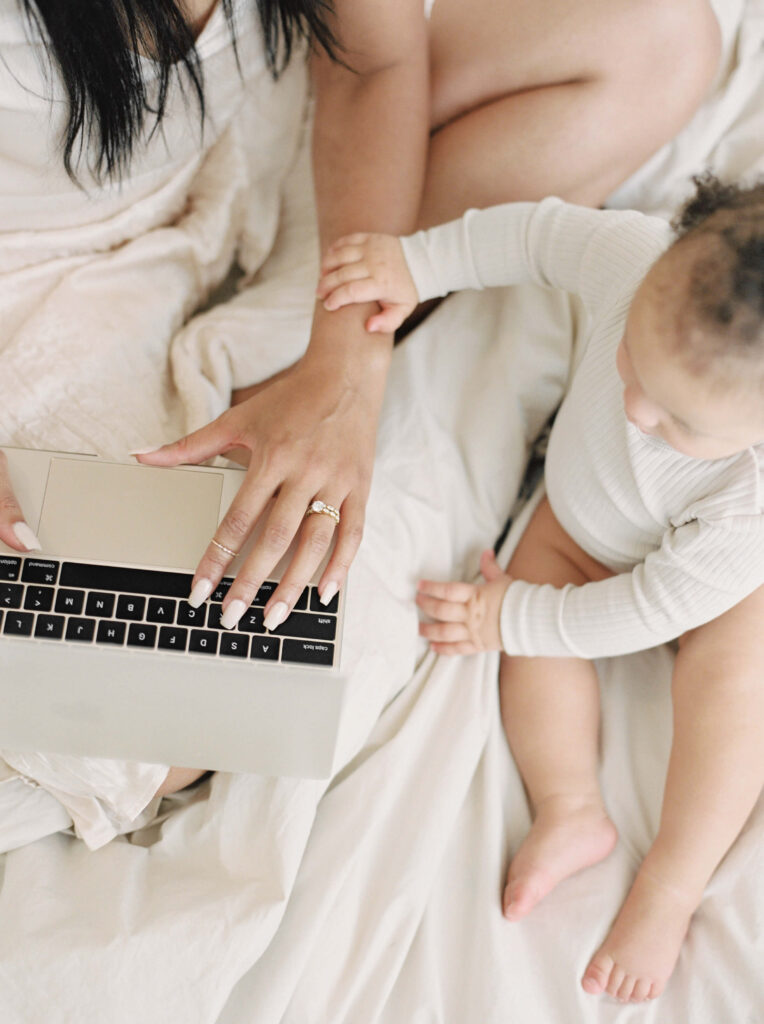 A mom working on her laptop while sitting in bed with her baby sitting next to her.